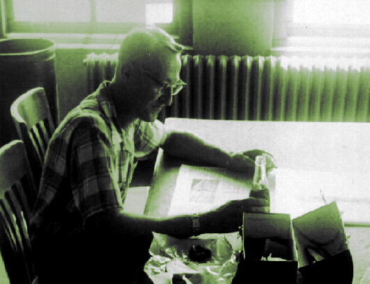Man 
sitting at a table with a newspaper, lunchbox and a cool drink