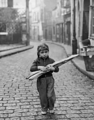 Small girl holding baguettes, 
standing in an empty, cobblestone street